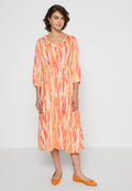 Langes, langärmliges Kleid im orange-rosa Batikmuster mit einer leichten Taillierung und V-Ausschnitt. Kombiniert mit orangefarbenen Ballerinas.