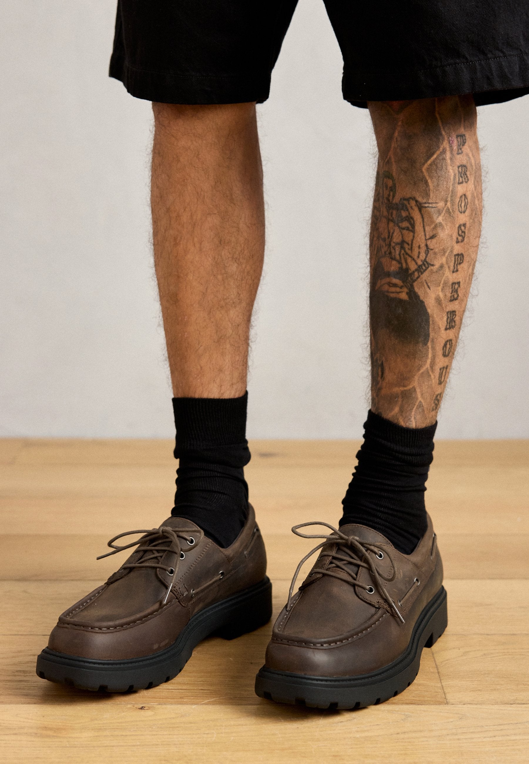 Braune Lederschuhe mit runden Zehen, schwarzen Gummisohlen und Schnürsenkeln. Getragen mit schwarzen Socken und Shorts, im Hintergrund ein Beintattoo.