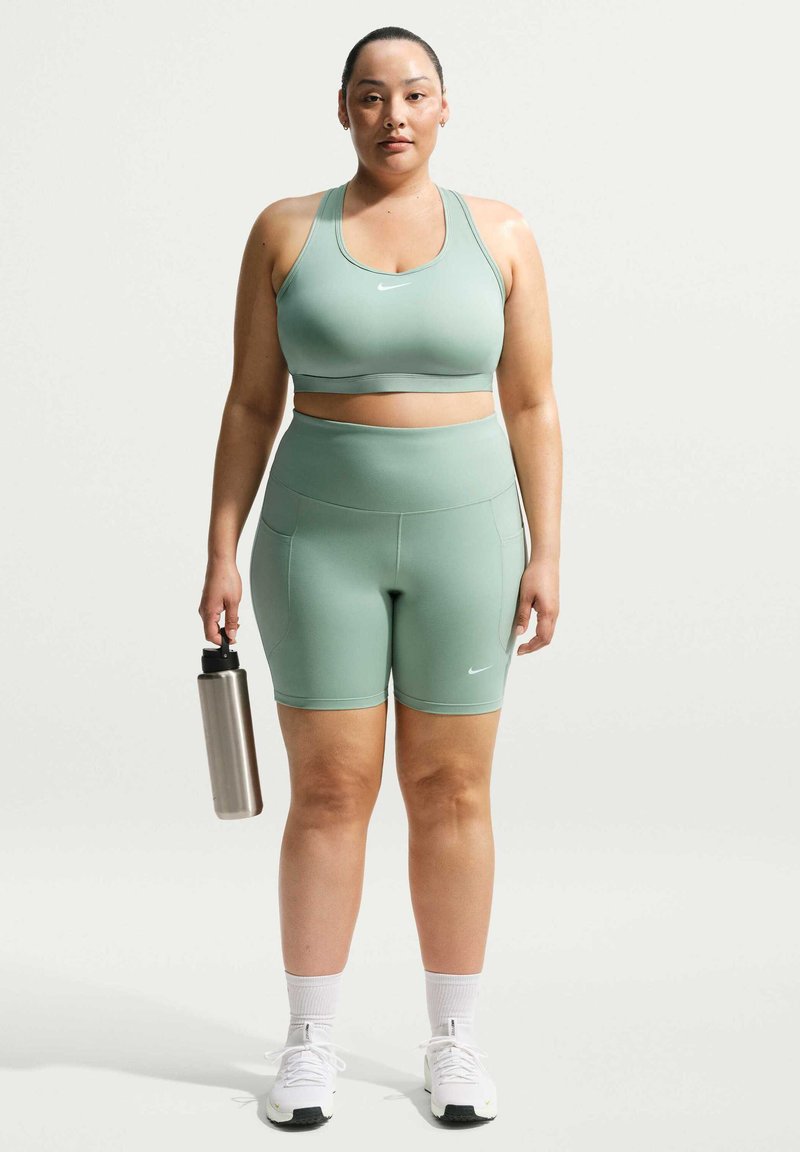 Frau in grünem Sport-BH und Shorts hält eine Edelstahl-Wasserflasche und steht vor einem schlichten hellen Hintergrund.