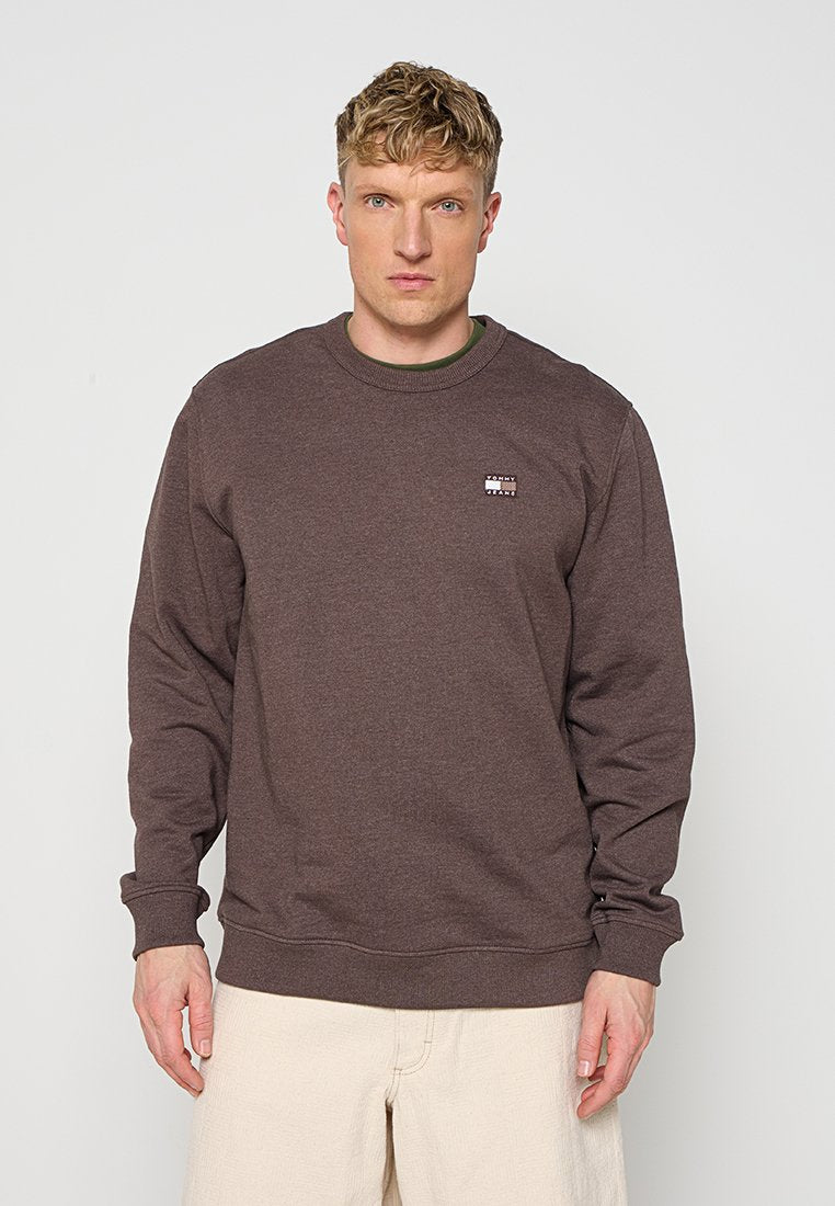 Mann mit kurzen blonden Haaren, der ein braunes langärmliges Sweatshirt mit Rundhalsausschnitt und hellbeige Hose vor einem schlichten Hintergrund trägt.