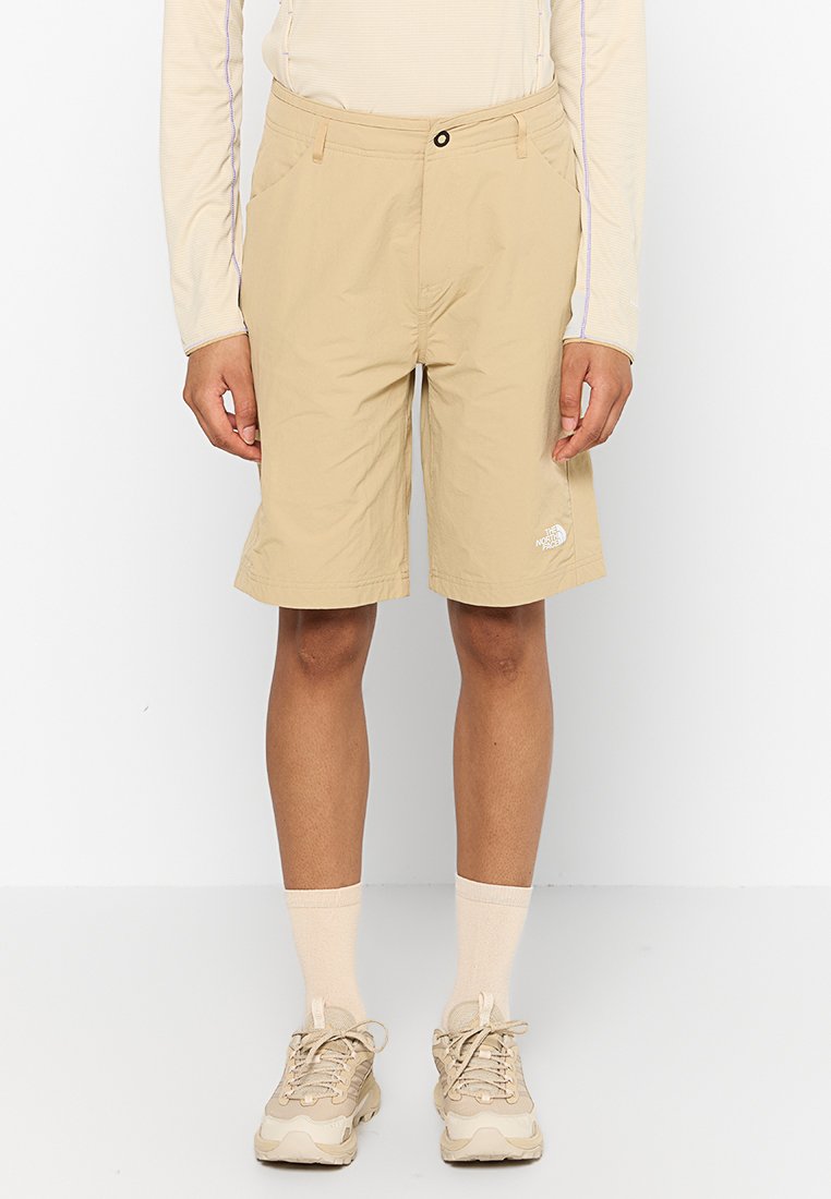 Beige Shorts aus leichtem Material, mit Reißverschluss, Seitentaschen und einem kleinen weißen Logo am linken Bein.