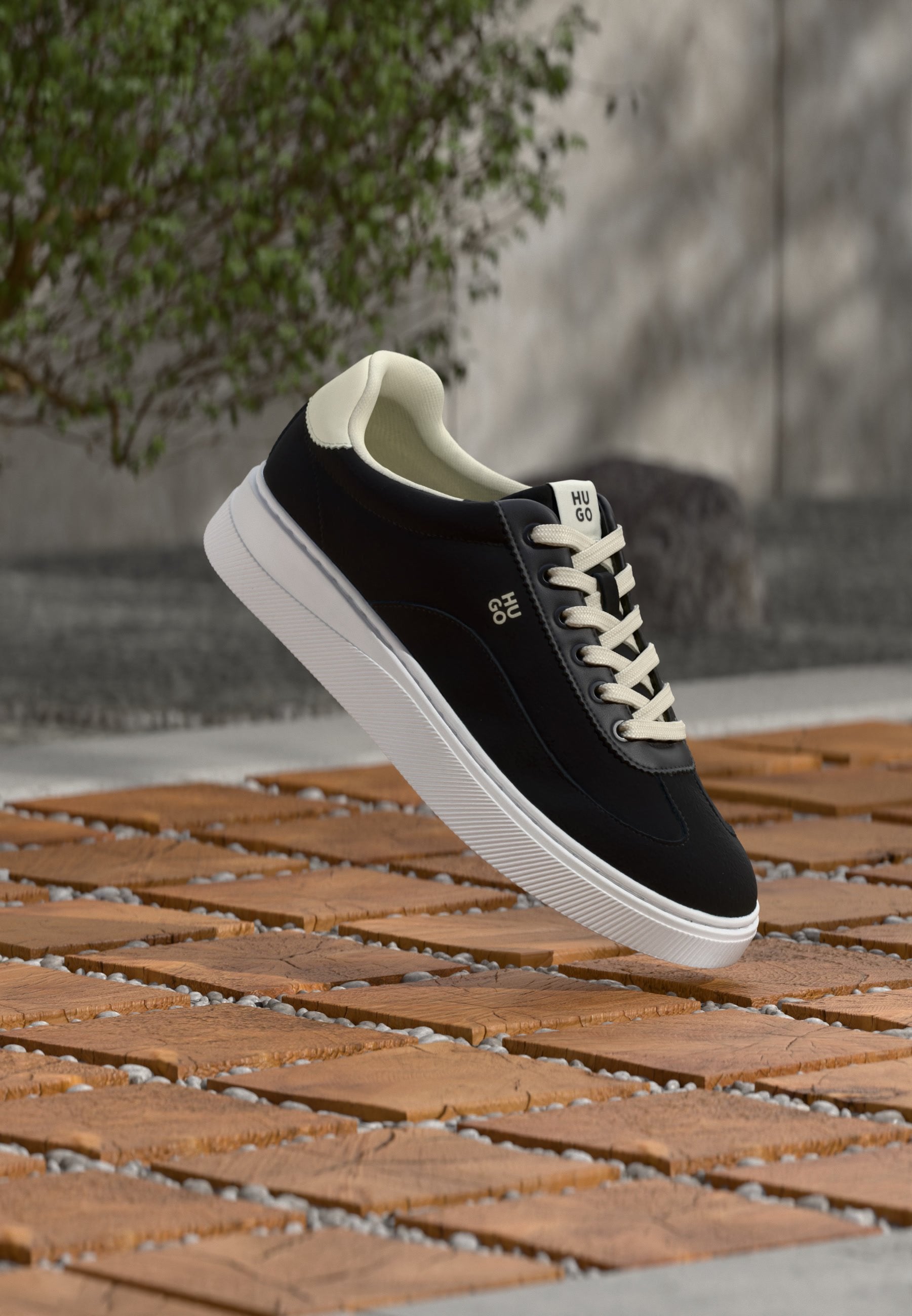 Schwarz-weißer Schnürsneaker mit weißer Sohle und "HUGO"-Logo, schwebt über einem gemusterten Untergrund aus Holz und Stein im Freien.