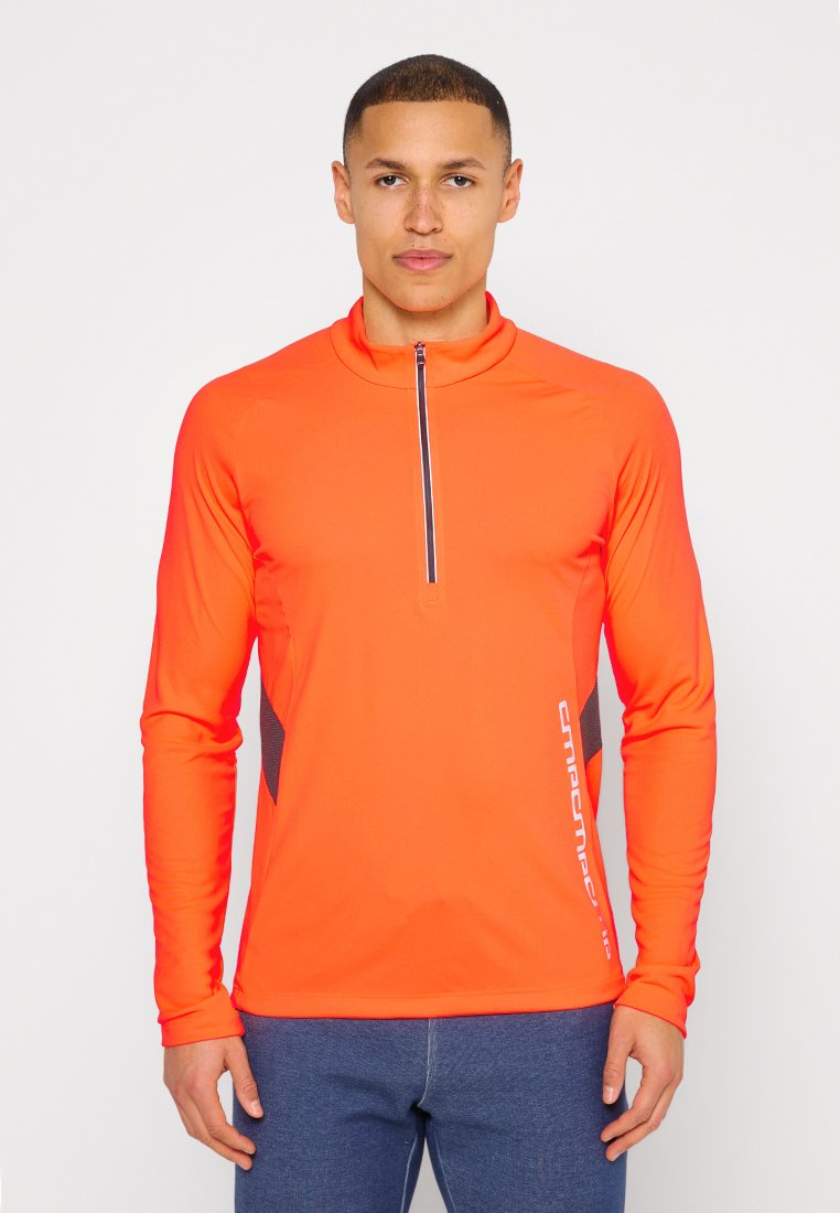 Mann trägt ein helloranges langärmliges Sportoberteil mit halbem Reißverschluss und dunkelblaue Hose, steht vor einem schlichten hellen Hintergrund.