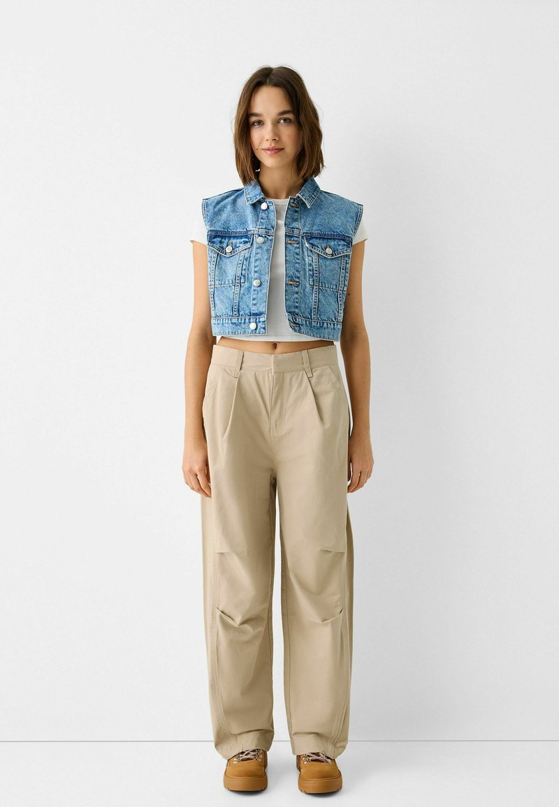 Denimweste mit ärmellosem Design, sanfter blauer Farbe und zwei Brusttaschen, kombiniert mit beigen, locker sitzenden Hosen und braunen Schuhen.
