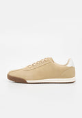 Beiger Sneaker mit flacher Silhouette, hellbrauner Sohle, weißem Fersen-Patch und beigen Schnürsenkeln auf schlichtem weißem Hintergrund.