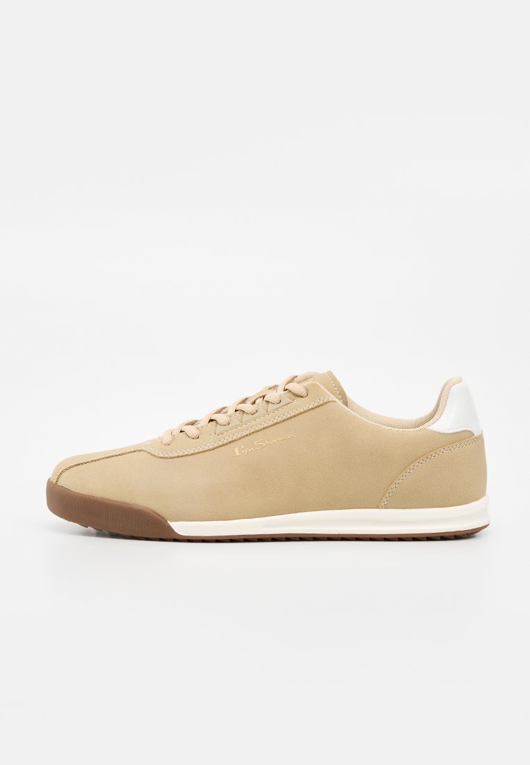 Beiger Sneaker mit flacher Silhouette, hellbrauner Sohle, weißem Fersen-Patch und beigen Schnürsenkeln auf schlichtem weißem Hintergrund.