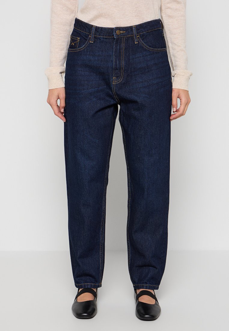 Dunkelblaue Jeans aus Denim mit geradem Schnitt, sichtbaren Nähten, fünf Taschen und einem Knopfverschluss, kombiniert mit schwarzen flachen Schuhen.