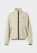 Beige Sherpa-Fleece-Jacke mit schwarzem Reißverschluss und Taschenbesatz, hohem Kragen, langen Ärmeln und einem kleinen rechteckigen Logo auf der Brust.