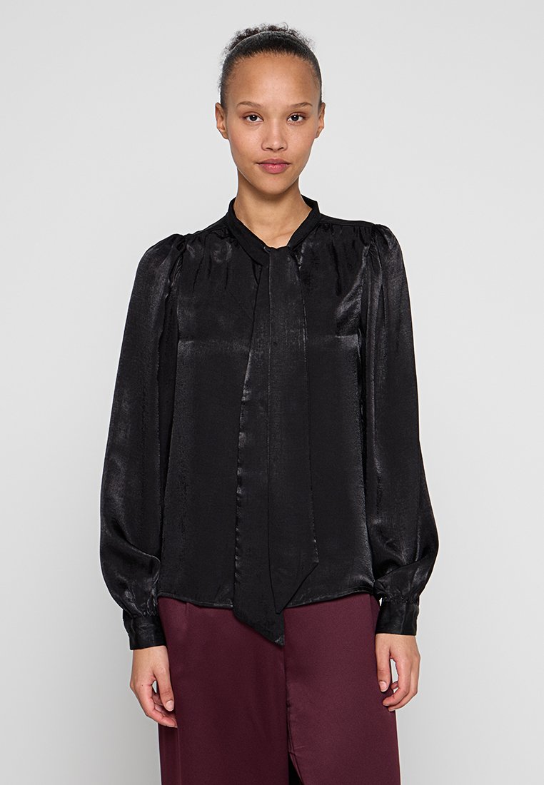 Schwarze Satinbluse mit langen Ärmeln, hohem Kragen und einer Bindung vorne. Glatte Textur und geraffte Schultern, getragen über bordeauxroten Hosen.