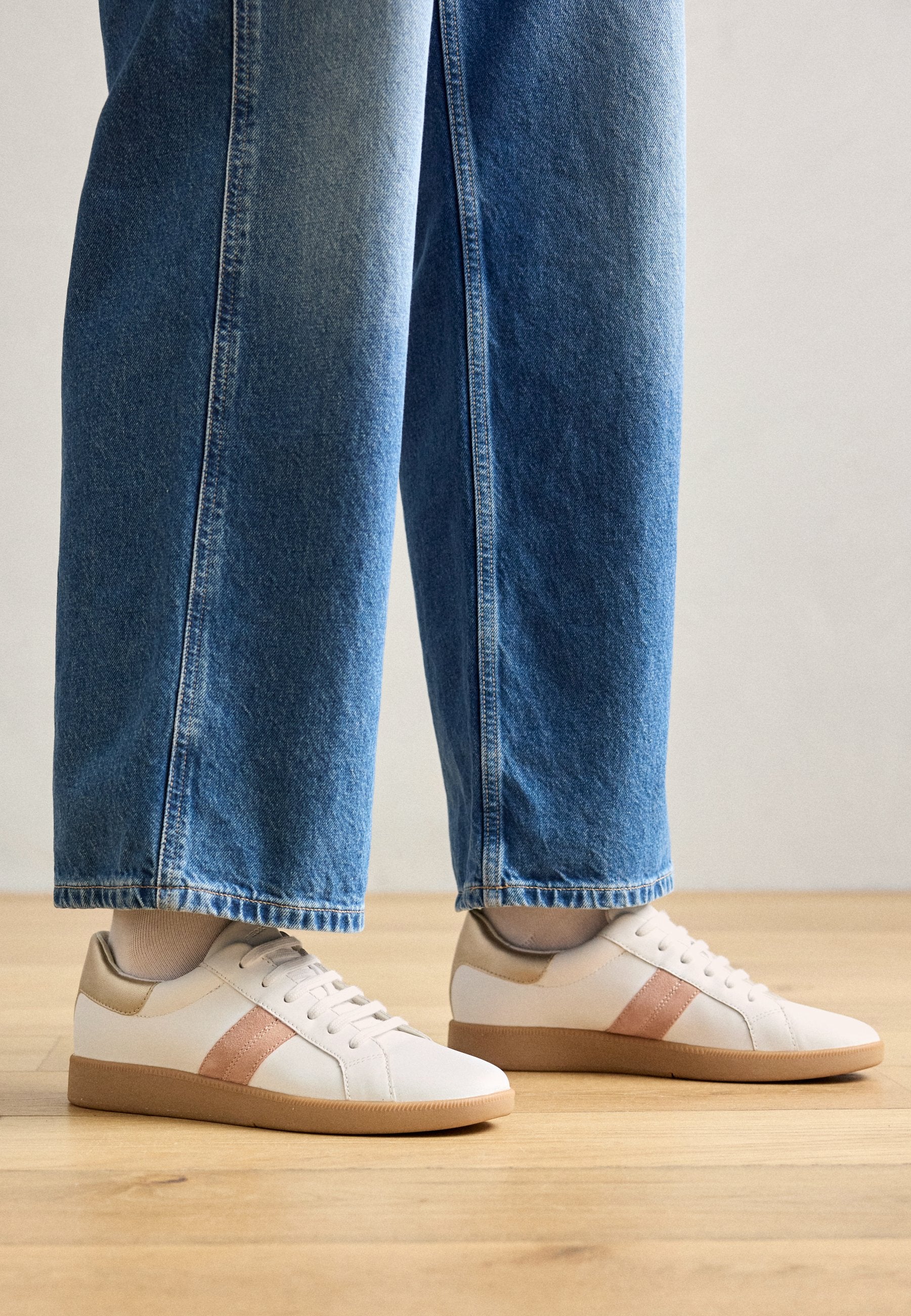Weiße Sneaker mit hellbraunen Akzenten, die eine glatte Textur und ein Schnürdesign aufweisen, sowie eine dicke beige Sohle haben, kombiniert mit blauen Jeans.