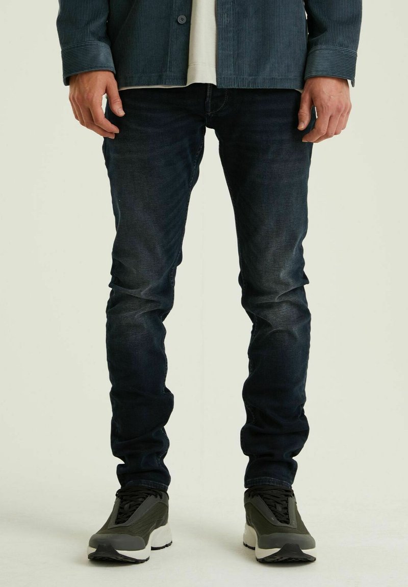 Dunkelblaue Slim-Fit-Jeans mit einem verblassten Design. Kombiniert mit grauen Sneakers, die eine auffällige weiße Sohle und schwarze Schnürdetails aufweisen.