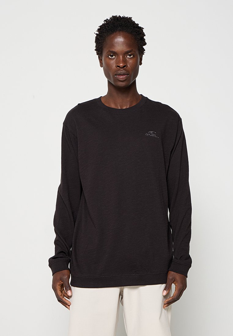 Mann trägt einen schwarzen O'Neill Sweatshirt mit langen Ärmeln und hellen Hosen und steht vor einem schlichten weißen Hintergrund.