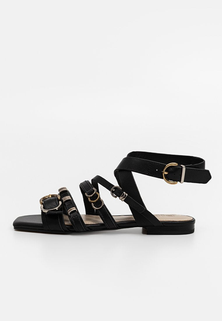 Schwarze flache Sandalen mit mehreren Riemen, die mit metallischen Schnallen und Ringen versehen sind. Hergestellt aus glattem Material, mit offener Zehenpartie und einem flachen Profilsohle.