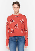 Roter Strickpullover mit abstrakten Mustern in Weiß und dunkelrot, Rundhalsausschnitt und verkürztem Design. Getragen mit blauen Jeans.