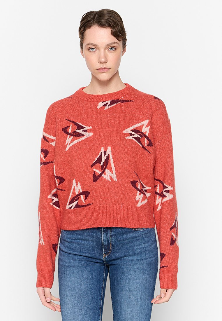 Roter Strickpullover mit abstrakten Mustern in Weiß und dunkelrot, Rundhalsausschnitt und verkürztem Design. Getragen mit blauen Jeans.