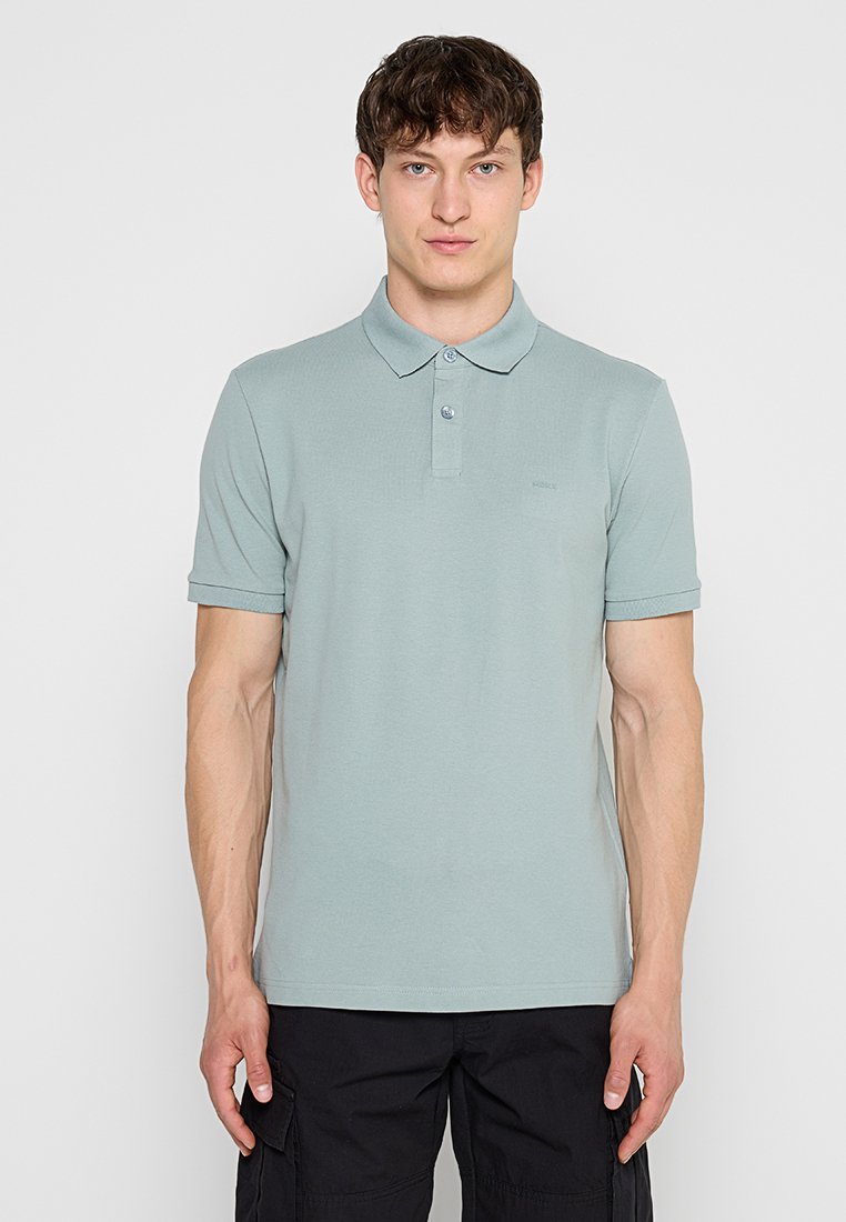 Junger Mann mit kurzen lockigen Haaren, der ein hellgrau-grünes Poloshirt und schwarze Cargohosen trägt, steht vor einem einfarbigen hellen Hintergrund.