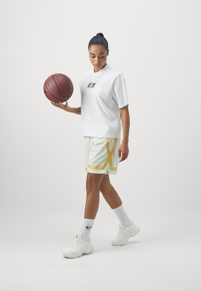 Frau in weißer Sportkleidung, die mit einer Hand einen Basketball hält und mit gesenktem Blick auf einem einfarbigen hellen Hintergrund geht.