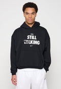 Junger Mann trägt einen schwarzen Hoodie mit dem Text "STILL TALKING" und kleinem Nike-Logo, kombiniert mit weißen Nike-Shorts.