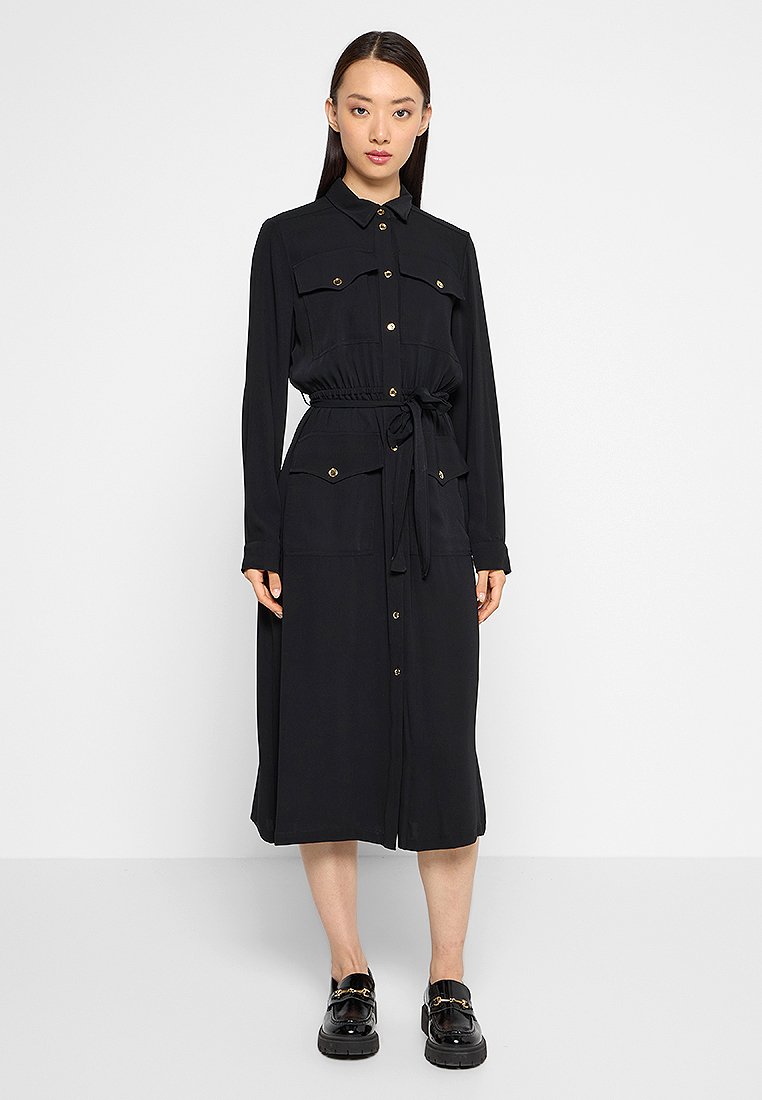 Schwarzes Hemdkleid mit langen Ärmeln, Knopfverschluss auf der Vorderseite, Brusttaschen und taillierter Taille. Hergestellt aus glattem Stoff und verfügt über goldfarbene Knöpfe.