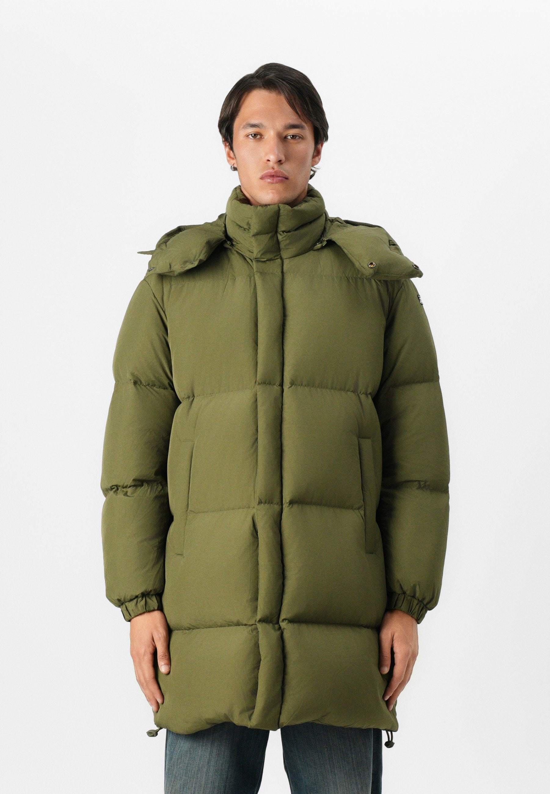 Mann trägt einen langen olivgrünen gepolsterten Wintermantel mit hohem Kragen und elastischen Bündchen und steht vor einem schlichten weißen Hintergrund.