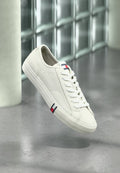 Weißer Tommy Hilfiger Sneaker mit Schnürsenkeln, der über einer grauen Oberfläche schwebt, im Hintergrund unscharf eine Glassteinwand und eine Säule.