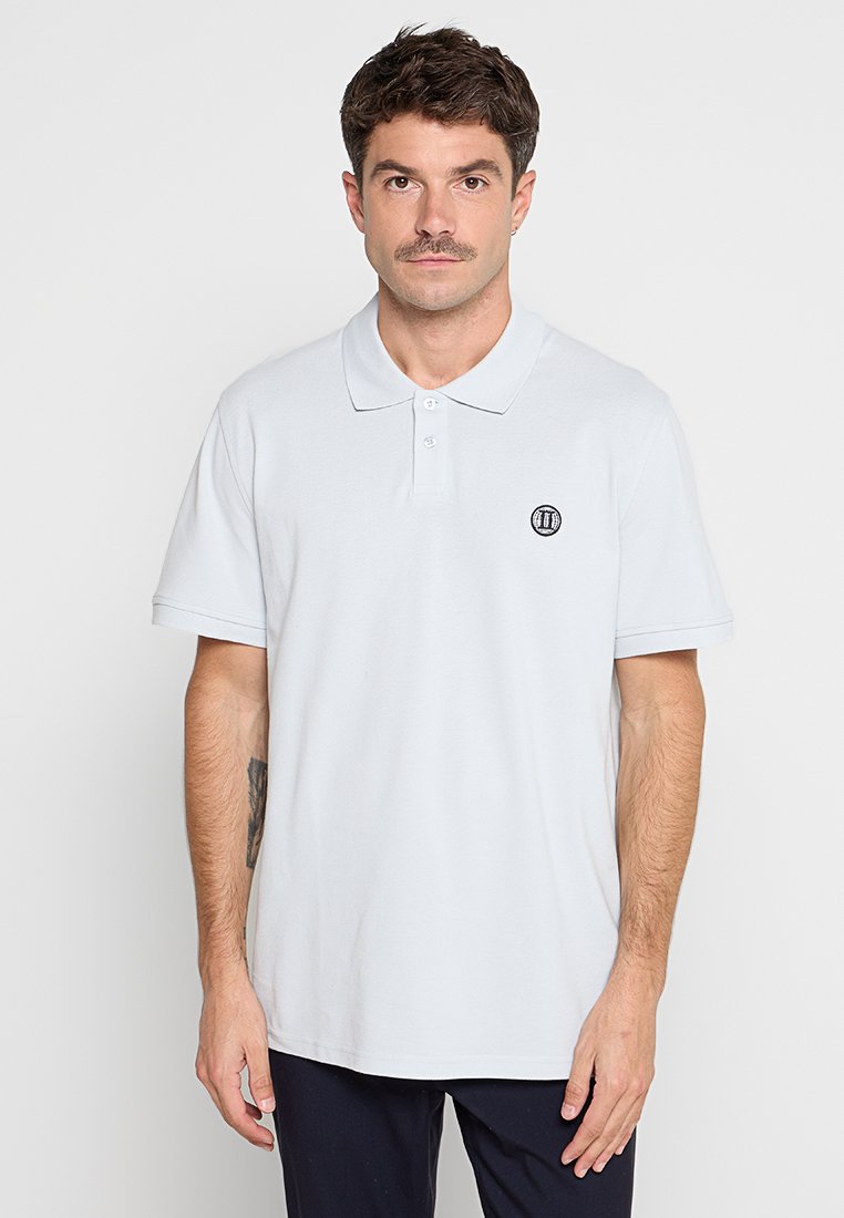 Mann mit kurzem braunem Haar und Schnurrbart trägt ein weißes Poloshirt mit aufgesticktem Logo auf der Brust und dunklen Hosen, steht vor einfarbigem Hintergrund.