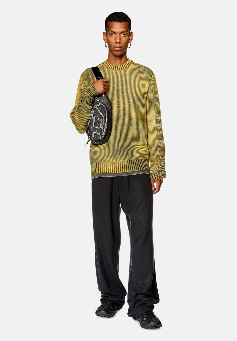 Gelber strukturierter Pullover mit Rippdesign, schwarze lässige Hose und eine schwarze Umhängetasche mit Grafik auf der Vorderseite.