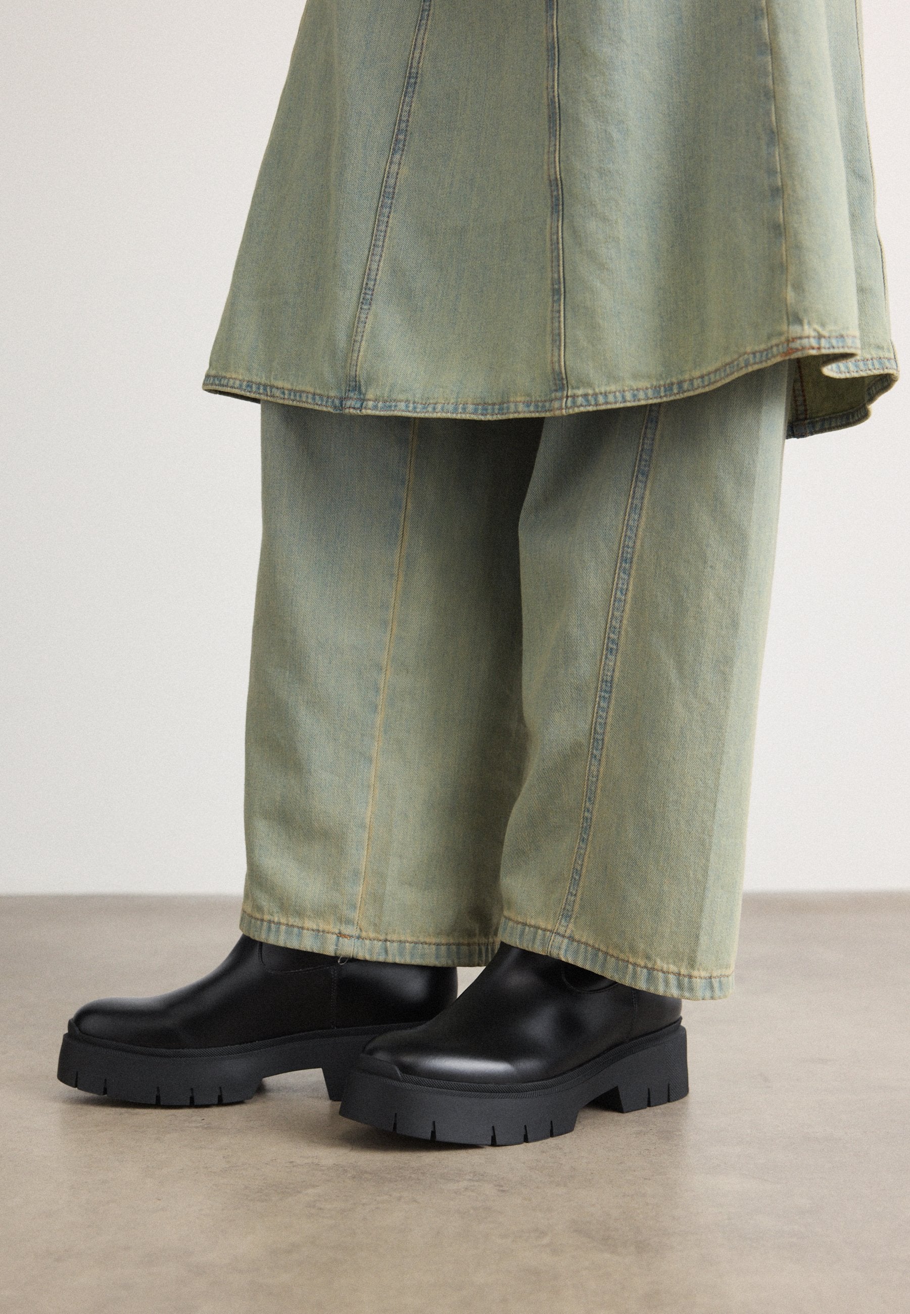 Schwarze Knöchelstiefel mit massiven, strukturierten Sohlen kombiniert mit weiten, hellgrünen Jeanshosen mit kontrastierenden Nähten.