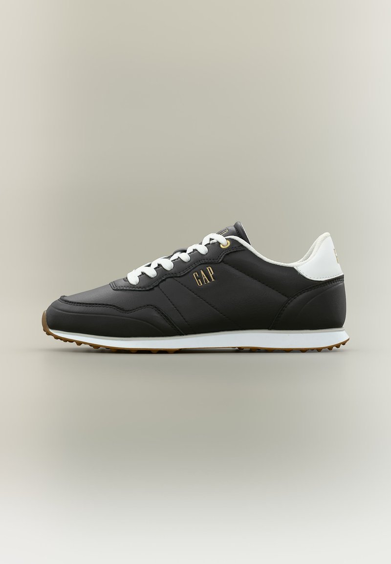 Schwarzer Sneaker in niedrigem Schnitt mit weißen Schnürsenkeln, weißem Fersenlaschen, brauner Sohle und goldenem "GAP"-Logo an der Seite vor einfachem Hintergrund.