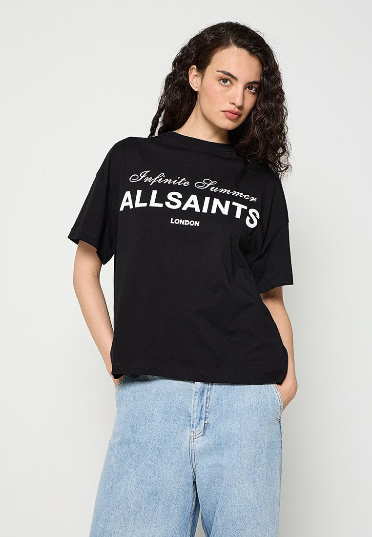 Junge Frau mit lockigem Haar, die ein schwarzes AllSaints T-Shirt mit dem Schriftzug „Infinite Summer“ und hellblaue Jeans trägt, Hände in den Taschen.