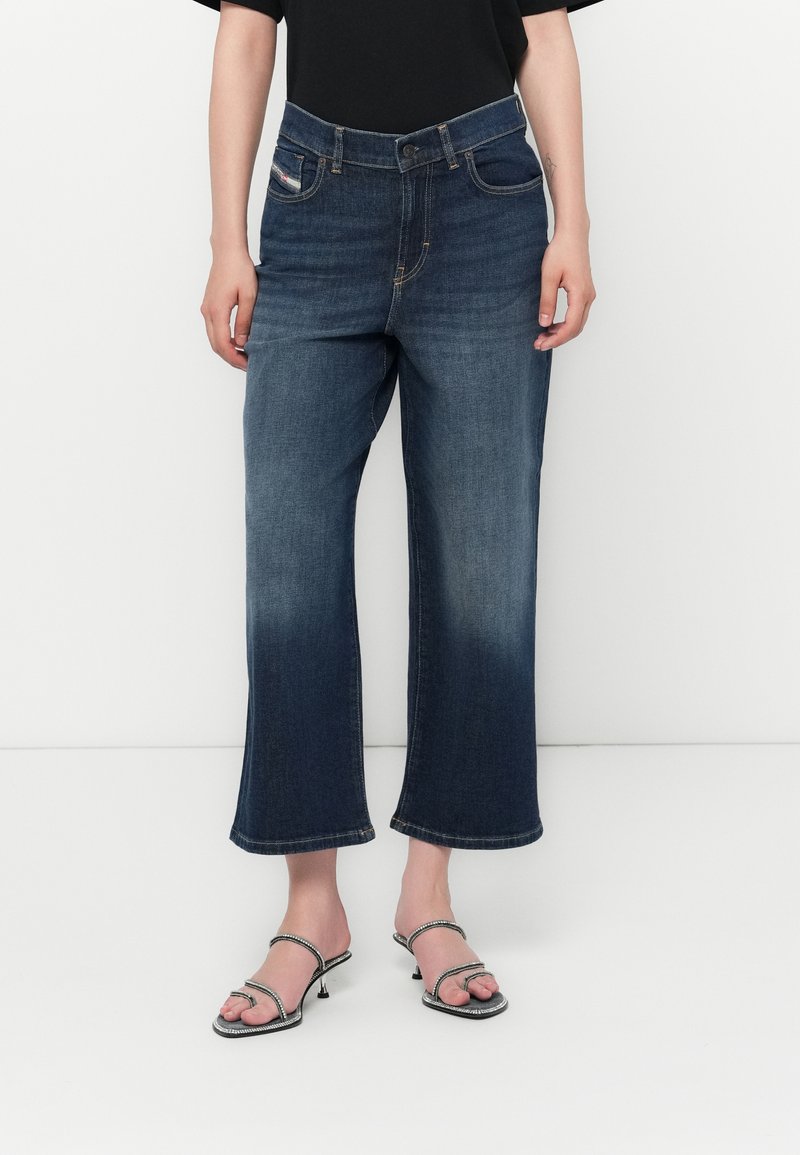 Dunkelblaue Jeans-Hosen mit weitem Bein und verblasstem Finish, ausgestattet mit Gürtelschlaufen, seitlichen Taschen und gesäumtem Saum. Getragen mit Riemchensandalen.