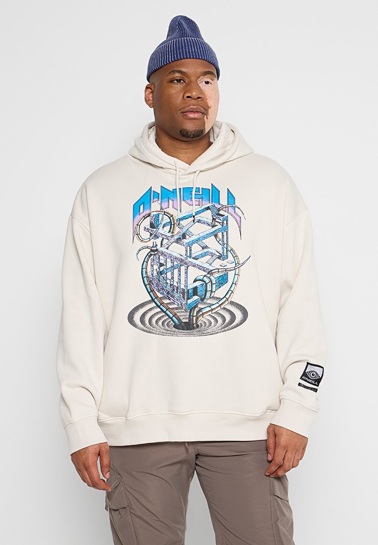 Mann mit einer Mütze, der einen beige Hoodie mit einem blauen abstrakten Würfedesign und braunen Cargo-Hosen trägt, steht vor einem schlichten Hintergrund.