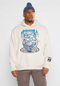 Mann mit einer Mütze, der einen beige Hoodie mit einem blauen abstrakten Würfedesign und braunen Cargo-Hosen trägt, steht vor einem schlichten Hintergrund.