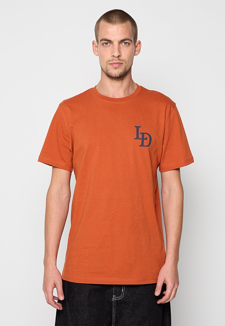 Oranges Baumwoll-T-Shirt mit kurzen Ärmeln, runder Halslinie und einem kleinen dunklen Logo im linken Brustbereich. Schlichte Textur, lockere Passform.