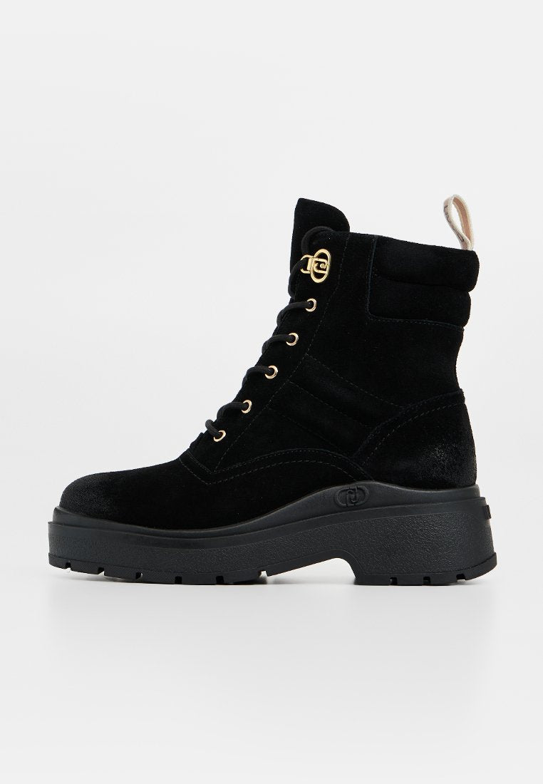 Schwarze Wildleder-Stiefeletten mit einer robusten schwarzen Sohle, goldenen Ösen und einem gepolsterten Schaft. Mit einer Schlaufe auf der Rückseite für einfaches Anziehen.