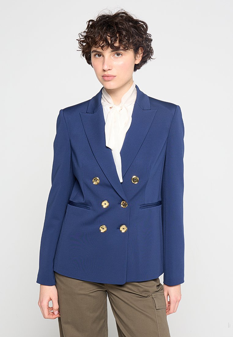 Marineblaues, doppelt verblasenes Blazer mit goldenen Knöpfen, scharfen Revers und zwei Seitentaschen, kombiniert mit einer weißen Bluse. Strukturierter Stoff.