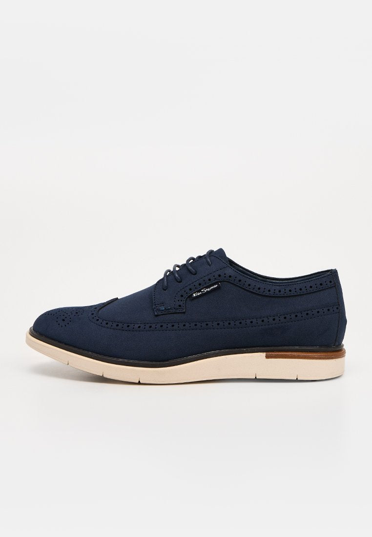 Marineblauer Herren-Brogue mit cremefarbener Sohle, Schnürverschluss, Flügelspitzen-Details und kleinem Markenetikett an der Seite.