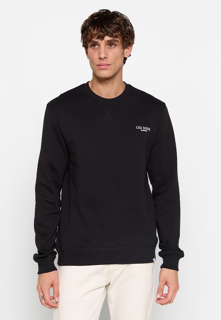 Schwarzer Hoodie mit geripptem Kragen, Bündchen und Saum. Besitzt ein kleines weißes Logo auf der Brust und einen strukturierten, gemusterten Stoff.