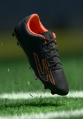 Schwarzer Fußballschuh mit orangefarbenen Akzenten, strukturiertem Obermaterial, Schnürsenkeln und Stollen. Er scheint über grünem Gras zu schweben.