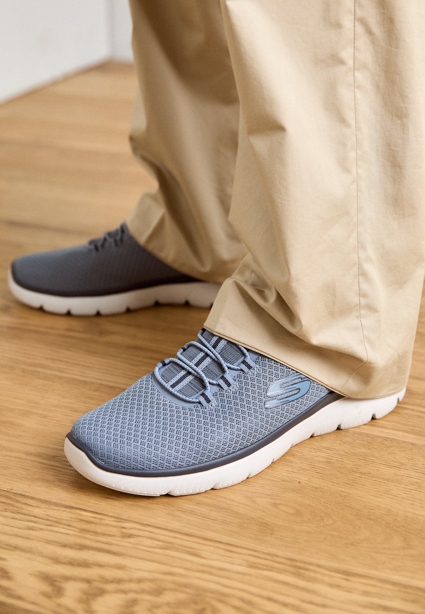 Helle graue Sportschuhe mit einem atmungsaktiven Mesh-Obermaterial, elastischem Schnürsystem, einer dicken weißen Sohle und einem Logo-Logo auf der Seite.