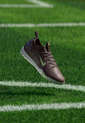 Dunkelbrauner Nike-Sneaker mit weißer Sohle und hellblauen Punkten, der über grünem Gras mit weißen Spielfeldlinien schwebt.