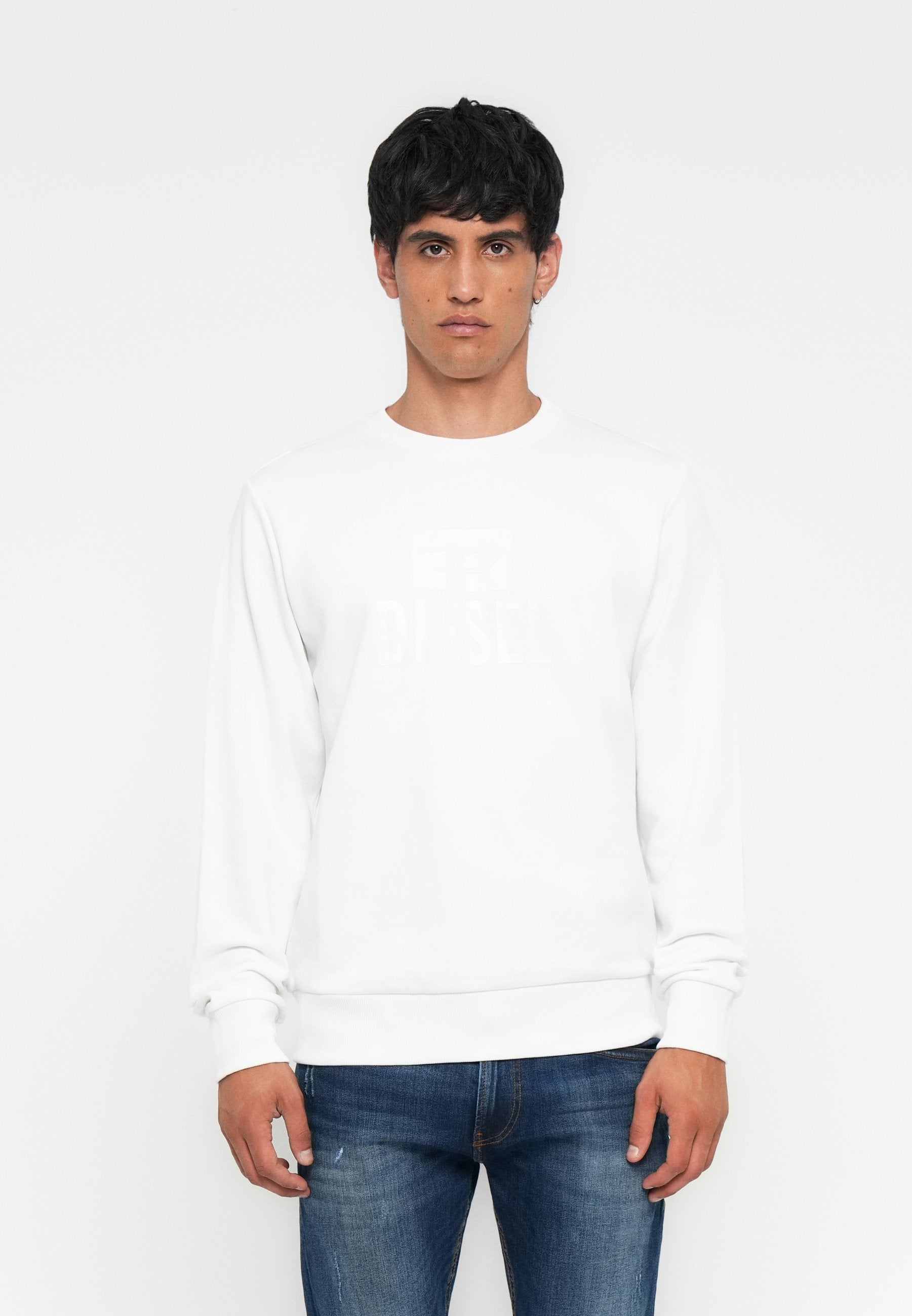 Mann in weißem langärmligen Diesel-Sweatshirt und blauer Jeans steht vor einem schlichten weißen Hintergrund, blickt nach vorne mit neutralem Ausdruck.