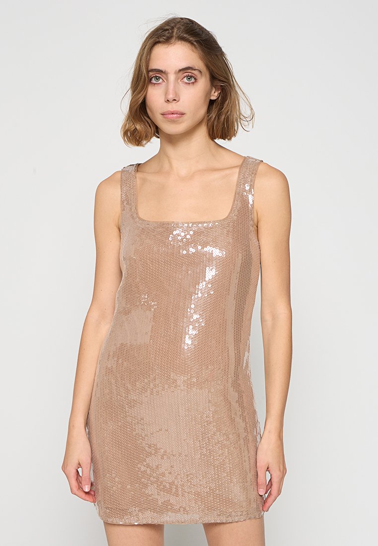 Junge Frau trägt ein ärmelloses, glitzerndes, beiges Mini-Kleid mit quadratischem Ausschnitt und steht vor einem schlichten hellgrauen Hintergrund.