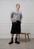 Kurzärmeliges Hemd mit floralem Monochrommuster, kombiniert mit schwarzen Shorts und schwarzen Knöchelboots. Vor einem minimalistischen Hintergrund.