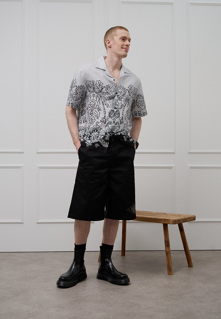 Kurzärmeliges Hemd mit floralem Monochrommuster, kombiniert mit schwarzen Shorts und schwarzen Knöchelboots. Vor einem minimalistischen Hintergrund.