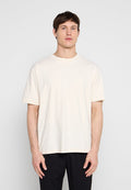 Junger Mann mit kurzen braunen Haaren, der ein schlichtes, hellbeiges, übergroßes T-Shirt und schwarze Hose trägt und vor einem schlichten grauen Hintergrund steht.