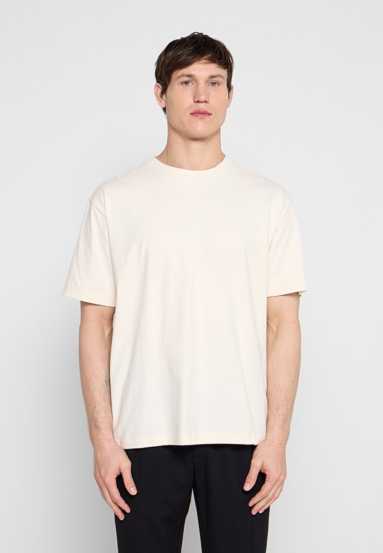 Junger Mann mit kurzen braunen Haaren, der ein schlichtes, hellbeiges, übergroßes T-Shirt und schwarze Hose trägt und vor einem schlichten grauen Hintergrund steht.