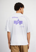 Mann mit kurzen, gedrehten Haaren, der ein weißes übergroßes T-Shirt mit lila Alpha Industries-Logo und Text auf der Rückseite trägt, kombiniert mit braunen Hosen.