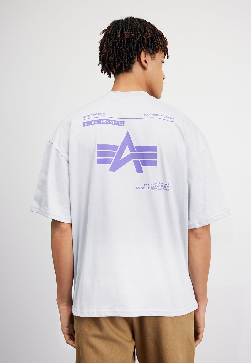 Mann mit kurzen, gedrehten Haaren, der ein weißes übergroßes T-Shirt mit lila Alpha Industries-Logo und Text auf der Rückseite trägt, kombiniert mit braunen Hosen.