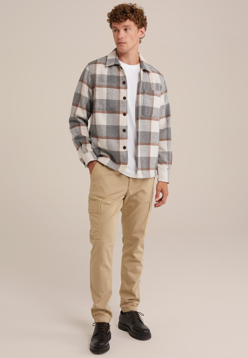 Junger Mann steht mit den Händen in den Taschen, trägt beige Cargo-Hosen, ein weißes T-Shirt, ein grau-braunes kariertes Hemd und schwarze Schnürschuhe vor einem einfarbigen Hintergrund.